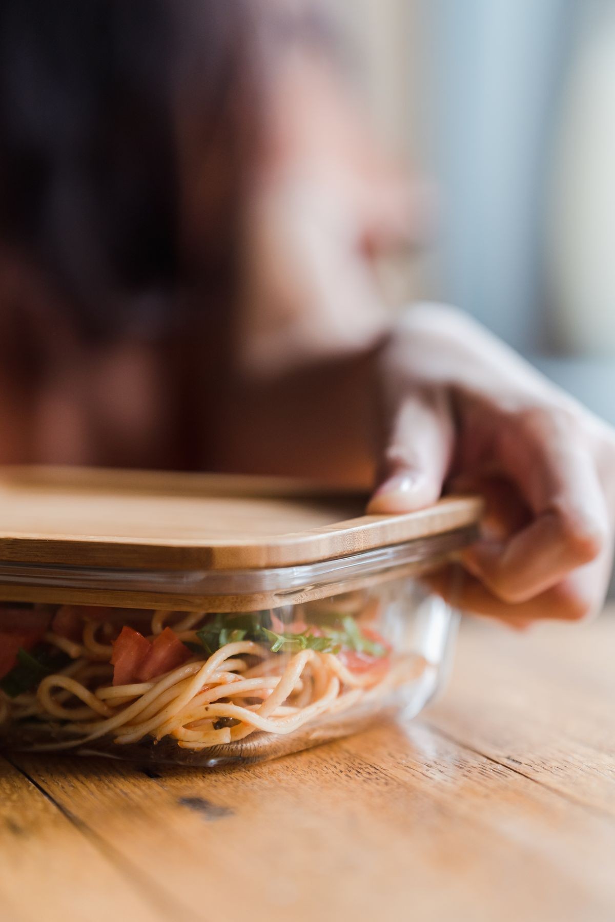 A hand places a wooden lid on a glass food container holding noodles
